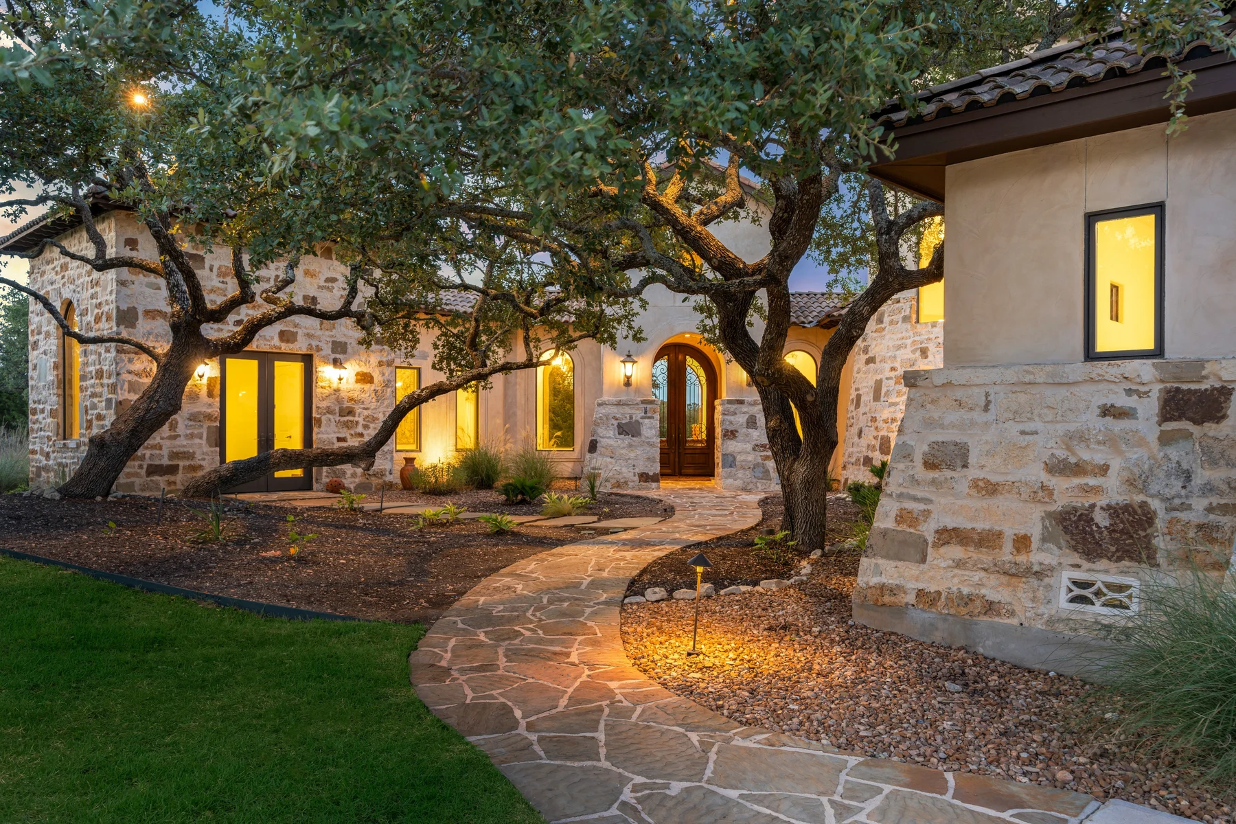 A tree-lined limestone home with an illuminated entry and a curving stone walkway, from Green Ease’s existing website.